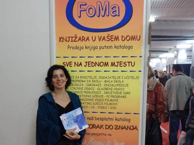 Interliber 2015., promocija slikovnice Krava koja je bila plava: sudionica sa slikovnicom ispred plakata izdavačke kuće FoMa